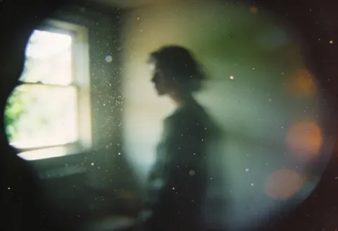 Dream behind glass — A softly blurred silhouette beside a bright window, photographed through misted glass with tiny light specks and rainbow reflections, quiet dreamlike mood.