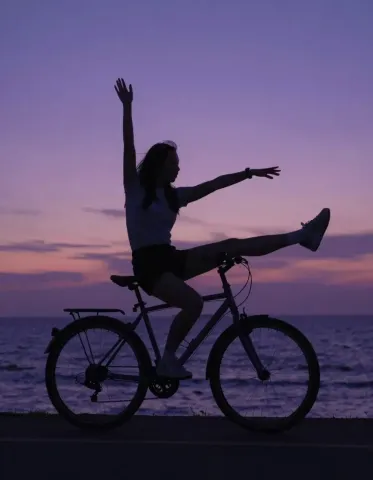Bicycle sunset — A playful cyclist silhouette beside the sea at purple sunset, outstretched arms and a clean horizon.