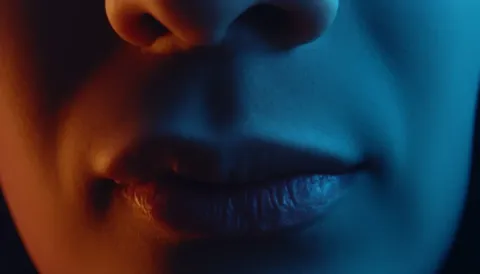 Split-light close-up — A tightly cropped face illuminated by orange and blue light, deep shadows and cinematic color contrast.